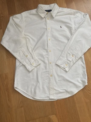 Vit skjorta från Polo Ralph Lauren - Klassisk vit skjorta från Polo Ralph Lauren med broderad logga på bröstet. Skjortan har lång ärm, knappar framtill och krage. Perfekt till jeans eller chinos för en clean och stilren look.