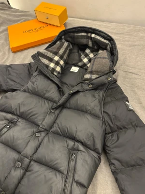 Svart dunväst med avtagbara ärmar av Burberry - Snygg svart dunväst från Burberry med avtagbara ärmar och huva. Västen har rutigt foder i huvan, två dragkedjefickor och knäppning framtill. Perfekt för lager-på-lager och håller dig varm under kyliga dagar.