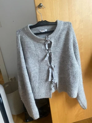 Grå stickad kofta med rosetter - Mysig grå stickad kofta med ribbad struktur och fyra dekorativa rosetter framtill istället för knappar. Koftan har rund halsringning och långa ärmar, perfekt för lager på lager och en söt touch till din outfit.