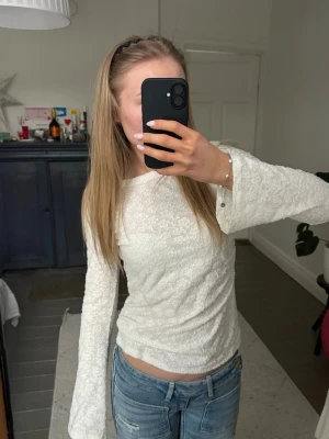 Vit långärmad spetsblus - Säljer en vit långärmad topp med blommig spets och lätt utsvängda ärmar. Toppen har en figurnära passform och rund halsringning. Perfekt att styla med jeans eller kjol för en trendig look.