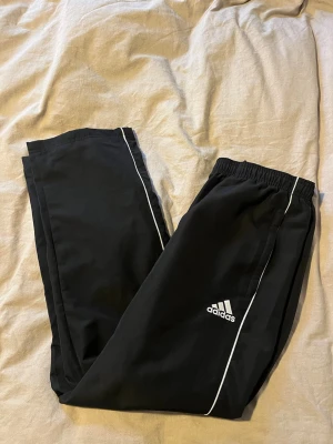 Svarta mjukisbyxor från Adidas - Svarta mjukisbyxor från Adidas med vita detaljer längs sidorna och klassisk logga på benet. Byxorna har elastisk midja och raka ben, perfekta för chill eller träning. Materialet känns lätt och sportigt.