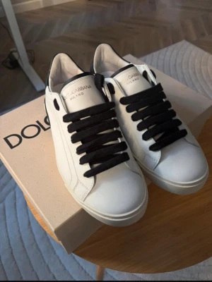 Dolce & Gabbana vita sneakers - Stilrena vita sneakers från Dolce & Gabbana med svarta detaljer och svarta snören. Skorna har rund tå, platt sula och logotyp på hälen. Tillverkade i skinn för en lyxig känsla och passar perfekt till en trendig streetwear-look.