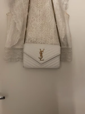Vit quiltad kuvertväska från YSL - Säljer en snygg vit kuvertväska från YSL med guldfärgad kedja och ikonisk YSL-logga framtill. Väskan är quiltad och har ett elegant kuvertformat lock. Perfekt accessoar för att lyfta vilken outfit som helst. Pris kan diskuteras för de som vill