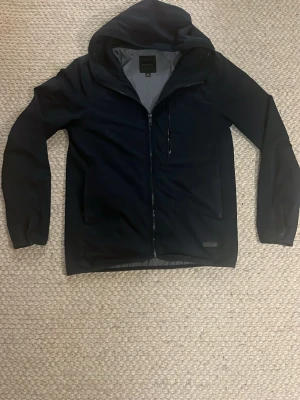 Svart windbreaker med huva från Elvine - Snygg svart windbreaker från Elvine i storlek XS. Jackan har huva, hel dragkedja framtill och praktiska fickor. Perfekt för blåsiga dagar och enkel att matcha med olika outfits. Stilren design och bekväm passform. Nypris 1200kr