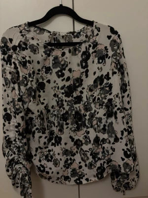 Mönstrad blus med knytärmar - Säljer en snygg blus med abstrakt svart, grått och beige mönster på vit botten. Blusen har långa ärmar med dekorativa knytband vid ärmsluten och rund halsringning. Materialet känns lätt och flowy, perfekt för en trendig look.