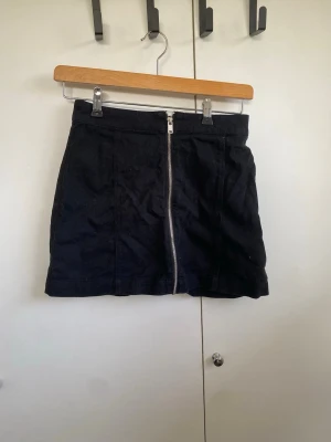 Svart kortkjol med dragkedja - Svart kortkjol i jeansmaterial med en snygg silverfärgad dragkedja. Kjolen har en rak passform och är enkel men stilren, perfekt att matcha med en cool t-shirt eller stickad tröja. Midjehög modell som ger en trendig look.