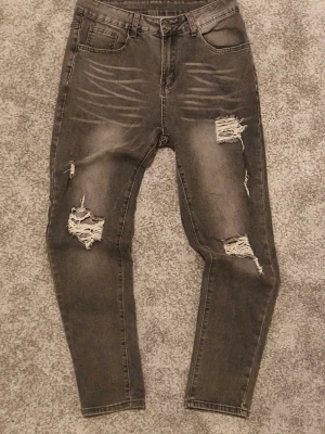 Grå slitna jeans med hål - Säljer ett par grå jeans med slitningar och stora hål på benen. Jeansen har fem fickor, dragkedja och knapp framtill samt en slim passform. Perfekta för en avslappnad och trendig look.