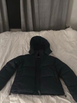 Mörkblå pufferjacka med huva - Snygg mörkblå pufferjacka med huva och dragkedja framtill. Jackan har en quiltad design och är vadderad för extra värme. Perfekt för kyliga dagar och har en klassisk, stilren look som passar till det mesta.