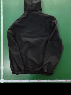 Svart vindjacka från Moncler - Snygg svart vindjacka från Moncler med huva och dragkedja framtill. Jackan har elastiska muddar vid ärmar och midja samt Moncler-logga på ena ärmen. Perfekt för blåsiga dagar och enkel att matcha med olika outfits.