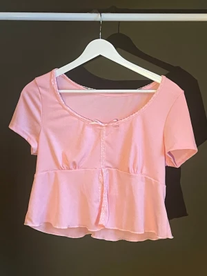 Ljusrosa croppad topp med volang - Söt ljusrosa croppad topp med korta ärmar och volang nedtill. Toppen har en liten rosett framtill, ribbad struktur och rundad halsringning med spetskant. Perfekt för en trendig och feminin look. Köpt från Sellpy, aldrig använt så helt nyskick🎀