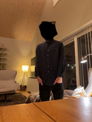 Mörkblå skjorta från Ralph Lauren - Klassisk mörkblå skjorta från Ralph Lauren med button-down krage och broderad logga på bröstet. Skjortan har långa ärmar och vita knappar framtill. Perfekt till jeans eller chinos för en clean och stilren look.