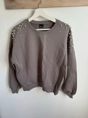 Grå sweatshirt med pärlor - Säljer en grå sweatshirt från Gina Tricot med dekorativa pärlor på axlarna. Tröjan har rund halsringning, långa ärmar och en avslappnad passform. Materialet är mjukt och bekvämt, perfekt för en trendig vardagslook. Dock finns en ljus fläck nertill, se bild 4.