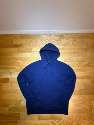 Mörkblå hoodie från H&M - En enkel och stilren mörkblå hoodie från H&M med klassisk huva och dragsnören. Hoodien har en stor magficka framtill och ribbade muddar vid ärmslut och nederkant. Perfekt för en avslappnad och casual look.