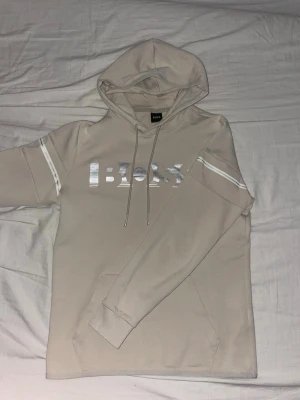 Beige/grå Boss hoodie - Snygg och stilren hoodie från HUGO BOSS i en elegant beige/off-white färg. Klassisk design med BOSS-logga i silver framtill samt diskreta reflexdetaljer på ärmarna som ger en modern look. Perfekt både till vardags och för en mer uppklädd streetstyle.  🔹 Skick: Nytt skick, sparsamt använd 🔹 Passform: Normal 🔹 Detaljer: Justerbar huva med snören 🔹 Material: Mjuk och bekväm kvalitet 🔹 Färg: Beige / ljus sand 🔹 Original HUGO BOSS  En tidlös hoodie som passar året runt