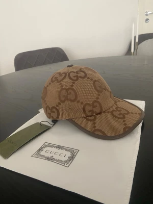 Brun keps från Gucci med GG-mönster - Säljer en snygg brun keps från Gucci med det klassiska GG-monogrammet i mörkare brunt över hela kepsen. Kepsen har böjd skärm och är tillverkad i ett slitstarkt tygmaterial. Perfekt för dig som vill ha en exklusiv och trendig accessoar.
