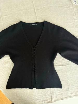 Svart Gina kofta  - Stilren svart kofta från Gina med v-ringning och dekorativa klädda knappar framtill. Figurnära passform och långa ärmar. Perfekt att matcha med jeans eller kjol för en trendig look.