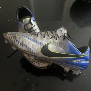 Nike Mercurial blå fotbollsskor - Säljer ett par Nike Mercurial fotbollsskor i blått och silver med svarta och gula detaljer. Skorna har snörning, platt sula och är tillverkade i syntetmaterial. Perfekta för dig som vill ha stilrena och snabba skor på planen.