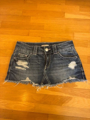 Blå mini kjol - Säljer en kort blå jeanskjol med slitna detaljer och fransig nederkant. Kjolen har klassiska fickor fram och bak samt bälteshällor. Perfekt för dig som gillar en avslappnad och trendig stil.