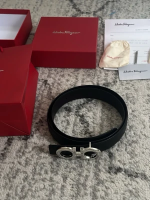 Svart Ferragamo bälte med silverdetalj - Snyggt svart bälte från Salvatore Ferragamo med ikoniskt silverfärgat spänne i metall. Bältet är tillverkat i skinn och har en stilren, klassisk design som passar till många olika outfits. Kommer med originalförpackning och dustbag.