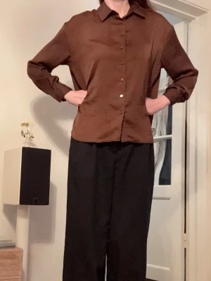 Brun Glansig Skjorta Stradivarius - Snygg brun skjorta med klassisk krage och knäppning framtill. Skjortan har lång ärm och en rak, lite loose passform. Perfekt för dig som gillar stilrena och enkla plagg med en modern touch. Material polyester.