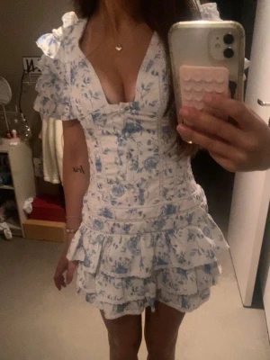 Blommig kortklänning med volanger - Supersöt kortklänning med blått blommönster på vit botten. Klänningen har volangdetaljer vid ärmar och kjol, v-ringning och smockad rygg för skön passform. Puffiga kortärmar och omlottkänsla framtill ger en romantisk vibe. Perfekt för sommarens alla tillfällen!