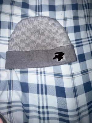 Grå mönstrad beanie med uppvik - Säljer en stilren grå beanie med diskret rutmönster och bred uppvikt kant. Mössan har en liten patch framtill och är gjord i mjukt stickat material som sitter skönt på huvudet. Perfekt för dig som vill ha en enkel men snygg accessoar till hösten och vintern.