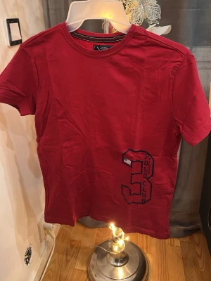 Röd t-shirt från Vinson Polo Club - Snygg röd t-shirt från Vinson Polo Club med stort tryck av siffran 3 och texten 'Polo Club' framtill. Klassisk rund halsringning och korta ärmar. Perfekt för en avslappnad och sportig stil.