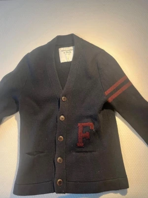 Mörkblå cardigan Abercrombie & Fitch - Collegeinspirerad mörkblå cardigan med knappar från Abercrombie & Fitch. Två röda ränder på ena ärmen och ett stort rött F i filt på bröstet. Djup V-ringning och två fickor framtill. Perfekt för en avslappnad men snygg stil.