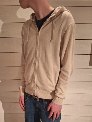Beige zip hoodie med huva - Snygg beige hoodie med dragkedja framtill och huva med dragsko. Tillverkad i mjukt, tunt material som känns skönt mot huden. Har ribbade muddar vid ärmslut och nederkant samt två fickor fram. Perfekt för en avslappnad och stilren look. Modelen är 172 cm!