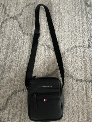 Svart axelväska Tommy Hilfiger - Snygg svart axelväska från Tommy Hilfiger i ett stilrent och kompakt format. Väskan har justerbar axelrem, en dragkedjeficka framtill och klassisk Tommy Hilfiger-logga i silver och färgdetalj. Tillverkad i slitstarkt skinnliknande material.