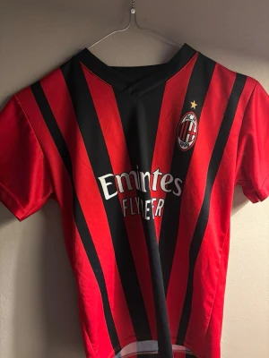 AC Milan matchtröja röd/svart - Säljer en klassisk AC Milan matchtröja i rött och svart med klubbmärke och Emirates Fly Emirates-tryck på bröstet. Tröjan har korta ärmar, V-ringning och är tillverkad i ett lätt och ventilerande material, perfekt för fotboll eller träning.