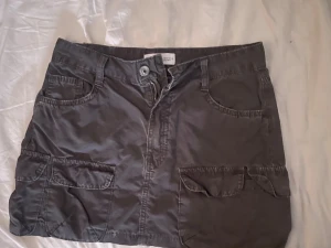 Grå kortkjol med fickor - Snygg grå kortkjol med flera stora fickor fram och bak samt bälteshällor. Kjolen har en rak passform och stängs med dragkedja och knapp framtill. Perfekt för en avslappnad och cool stil.