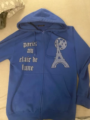Blå hoodie med Eiffeltornet och text - Snygg blå hoodie med dragkedja från BLINGLUCID. På framsidan finns ett stort tryck av Eiffeltornet och månen i glittriga stenar samt texten 'paris au clair de lune' i gotisk stil. Hoodien har huva med dragsko och två fickor framtill.