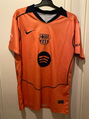 Fc Barcelona orange matchtröja Nike - Orange t-shirt från Nike med FC Barcelona-logga och Spotify-tryck på bröstet. T-shirten har mörkblå detaljer vid kragen och axlarna samt UNHCR-tryck på ryggen. Tillverkad i lätt polyester, perfekt för dig som gillar sportig stil.