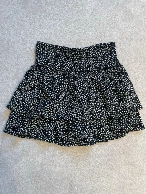 Svart blommig volangkjol med shorts under - Supersöt svart kortkjol med vitt blommönster och volanger. Kjolen har en bred resår i midjan för skön passform och är gjord i ett lätt, luftigt material. Perfekt för dig som vill ha en trendig och bekväm kjol med extra volym. Med inbyggda shorts under.