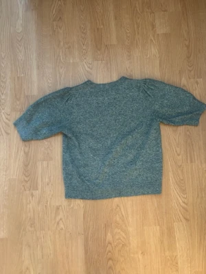 Grönmelerad stickad tröja - Mysig grönmelerad stickad tröja med rund hals och halvlånga, lätt puffade ärmar. Tröjan har en avslappnad passform och ribbstickade muddar vid ärmslut och nederkant. Perfekt för lager på lager under kyliga dagar. Storlek small.