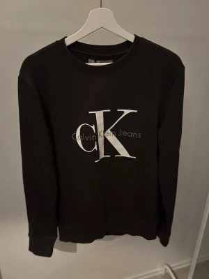 Svart sweatshirt från Calvin Klein Jeans - Stilren svart sweatshirt från Calvin Klein Jeans med stort vitt CK-tryck på bröstet. Tröjan har rund halsringning, långa ärmar och ribbade muddar vid ärmslut och nederkant. Perfekt för en avslappnad och trendig look.