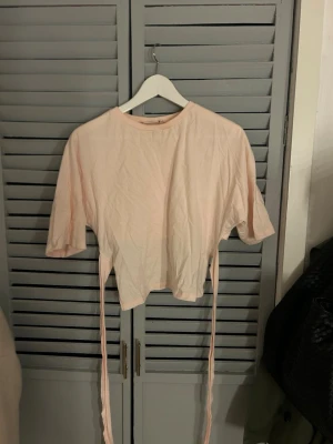 Ljusrosa croppad topp med knyt - Säljer en ljusrosa croppad topp med korta ärmar och långa band som knyts i ryggen för en snygg detalj. Toppen har enkel design och är perfekt att styla med höga jeans eller kjol. Materialet känns mjukt och lätt.