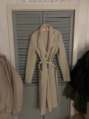 Beige kappa med bälte och krage - Stilren beige kappa med bred krage och knytskärp i midjan. Långärmad modell med klassisk passform och snyggt fall. Perfekt för dig som vill ha en enkel men elegant look till höst och vinter.