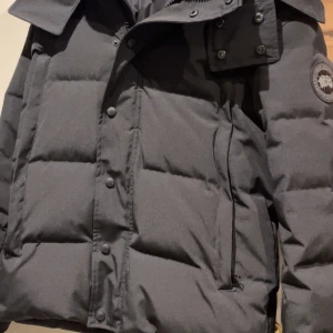 Svart wyndham från Canada Goose - Säljer en svart dunjacka från Canada Goose med klassisk patch på ärmen. Jackan är quiltad, har dragkedja och knappar framtill samt hög krage. Perfekt för kalla vinterdagar och har en stilren, mörk look.