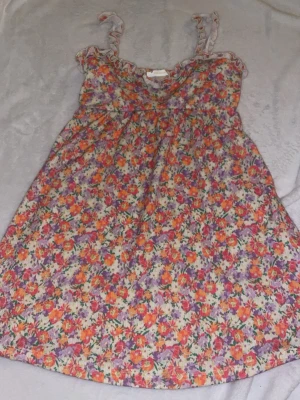 Blommig sommarklänning med volang - Söt kort blommig klänning med volangkant och smala axelband. Färgglatt mönster i lila, orange och rött på ljus botten. Perfekt för varma dagar och har en luftig passform. 