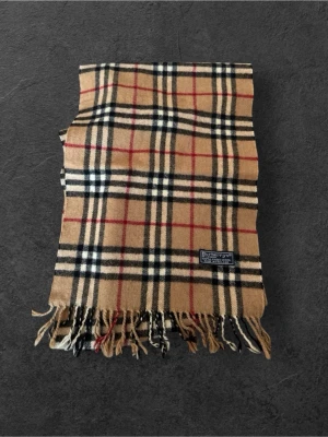 Beige rutig halsduk från Burberry - Klassisk beige halsduk från Burberry med det ikoniska rutmönstret i svart, vitt och rött. Halsduken har fransar i ändarna och är tillverkad i mjuk ull som känns lyxig och varm. Perfekt accessoar för att lyfta din outfit.