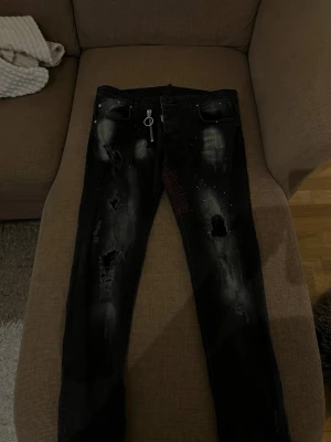 Svarta Dsquared2 jeans med dragkedja - Säljer ett par svarta jeans från Dsquared2 med slitningar, hål och coola detaljer. Jeansen har en stor silverfärgad dragkedja framtill och vita färgstänk för en edgy look. Passar perfekt för dig som gillar streetstyle och vill sticka ut.
