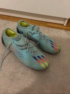Blåa Adidas fotbollsskor med snörning - Säljer ett par ljusblåa Adidas fotbollsskor med snörning och färgglada detaljer i rött, blått och gult. Skorna har en låg profil och ovandel i syntetmaterial med mönstrad textur för bättre bollkontroll. Perfekta för dig som vill sticka ut på planen.