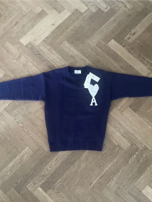 Mörkblå stickad tröja från Ami - Snygg mörkblå stickad tröja från Ami med rund halsringning och ribbade muddar. På bröstet finns ett vitt hjärta och bokstaven A, vilket är Ami:s ikoniska logga. Perfekt för dig som gillar stilrena plagg med en twist.