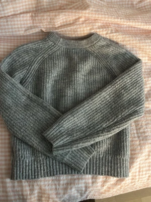 Grå stickad tröja från Lindex - Mysig grå stickad tröja från Lindex med ribbstickade muddar och rund hals. Tröjan har en klassisk rak passform och är perfekt för kyliga dagar. Tillverkad i mjukt material som känns skönt mot huden.