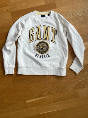 Vit sweatshirt från GANT - Vit sweatshirt från GANT med stor gul och svart logotyp och texten 'MCMLXIX' framtill. Tröjan har rund halsringning, ribbade muddar vid ärmslut och nederkant samt en avslappnad passform. Perfekt för en sportig och casual look.
