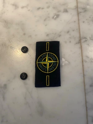 Stone Island badge med knappar - Säljer en klassisk svart Stone Island badge med broderad kompasslogga i gult och grönt. Två tillhörande svarta knappar ingår för enkel fastsättning på jacka eller tröja. Perfekt detalj för att ge din outfit en exklusiv touch. En kostar 30kr alla kostar 120