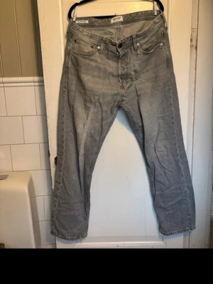 Jack & Jones ljusgrå jeans - Ljusgrå jeans med klassisk rak passform och femficksmodell. Modell Relaxed/Chris. Jeansen har knappgylf och är tillverkade i slitstark bomullsdenim. Perfekta för dig som gillar en tidlös och avslappnad look. Storlek 32/30