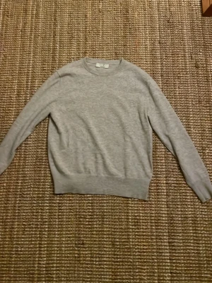 Ljusgrå stickad tröja från Carmes - En stilren ljusgrå stickad tröja från Carmes med rund halsringning och långa ärmar. Tröjan har ribbstickade muddar vid ärmslut och nederkant, perfekt för lager-på-lager och enkel att matcha med olika outfits.